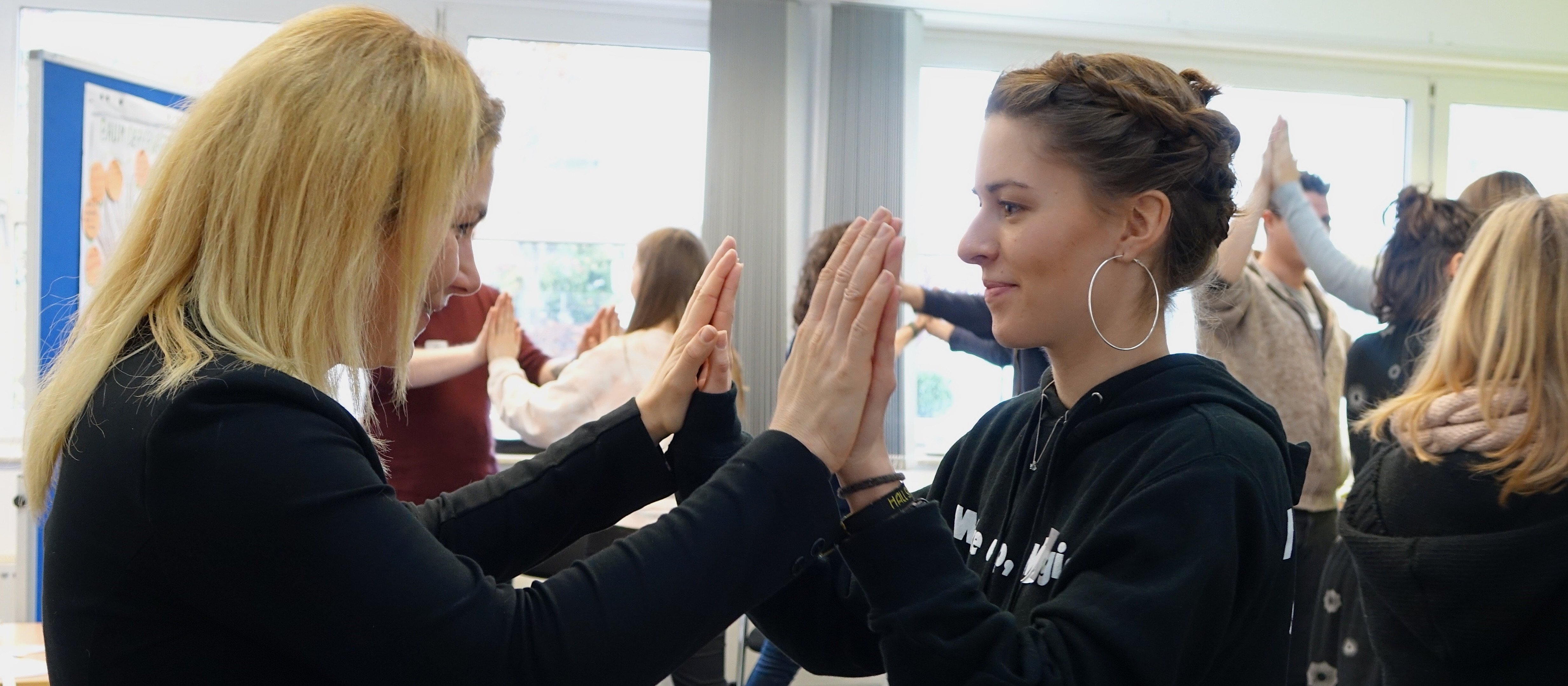 KI generiert: Das Bild zeigt zwei Frauen in einem Raum mit anderen Personen im Hintergrund, die sich gegenseitig die Handflächen zuwenden, als würden sie eine Übung oder ein Spiel durchführen. Der Hauptinhalt des Bildes ist die Interaktion zwischen den beiden Frauen im Vordergrund.