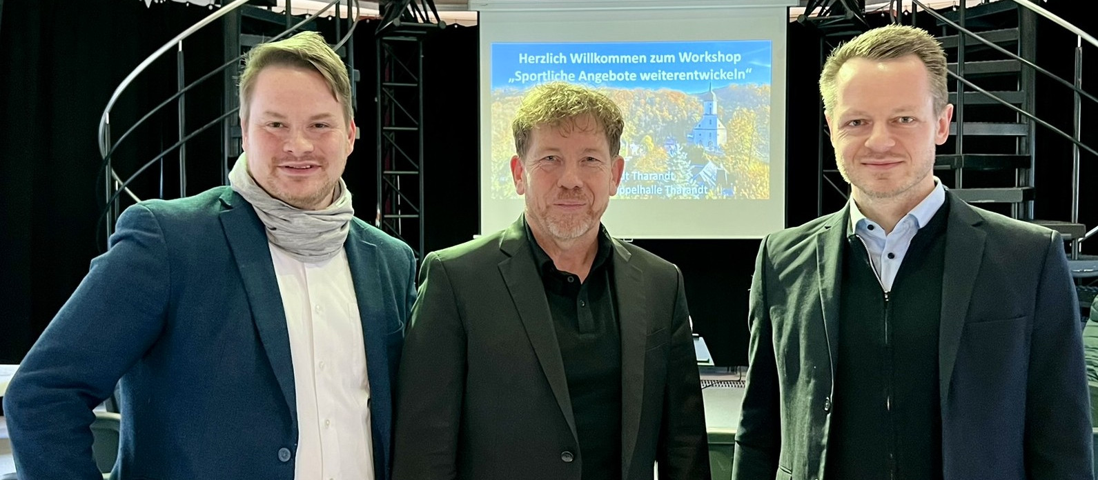 KI generiert: Das Bild zeigt drei Männer in Businesskleidung, die vor einer Leinwand mit der Aufschrift "Herzlich Willkommen zum Workshop 'Sportliche Angebote weiterentwickeln'" stehen. Der Hauptinhalt des Bildes ist die Präsentation oder der Workshop zu sportlichen Angeboten.