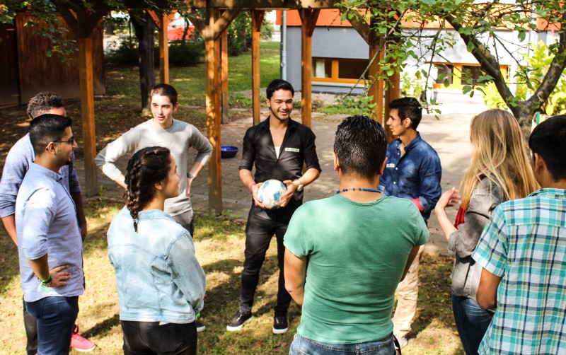 KI generiert: Das Bild zeigt eine Gruppe von Menschen, die im Freien in einem Kreis stehen und sich unterhalten, wobei eine Person in der Mitte einen Fußball hält. Der Hauptinhalt des Bildes ist eine informelle Zusammenkunft oder Diskussion in einer entspannten, natürlichen Umgebung.