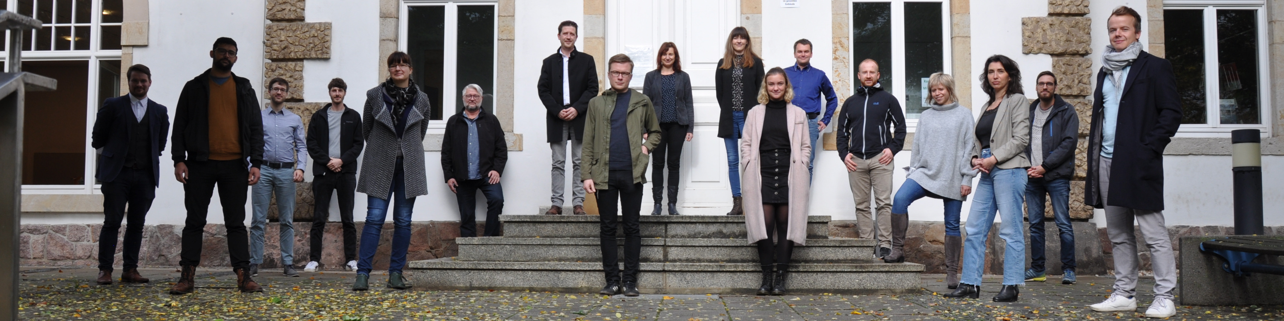 KI generiert: Das Bild zeigt eine Gruppe von 17 Personen, die vor einem Gebäude mit großen Fenstern und einer Treppe posieren. Die Personen sind in verschiedenen Kleidungsstilen gekleidet und stehen in einer lockeren Formation auf einem mit Blättern bedeckten Boden.