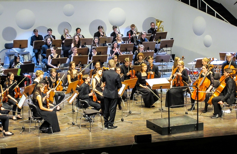 KI generiert: Das Bild zeigt ein Orchester, das auf einer Bühne in einem modernen Konzertsaal spielt. Im Vordergrund sind die Streicher zu sehen, während im Hintergrund die Bläser und Schlagzeuger positioniert sind.