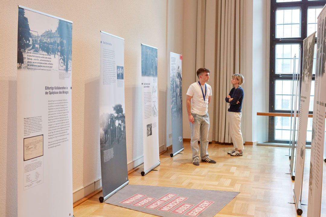 KI generiert: Das Bild zeigt eine Ausstellung in einem hellen Raum mit mehreren Informationsständern, die historische Fotos und Texte präsentieren. Zwei Personen, ein Mann und eine Frau, betrachten die Ausstellungsstücke und scheinen sich darüber auszutauschen.