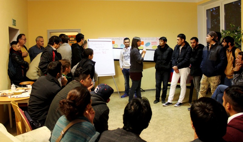 KI generiert: Das Bild zeigt eine Gruppe von Personen in einem Raum, wobei einige stehend und andere sitzend sind. Im Vordergrund präsentieren zwei Personen Inhalte auf Flipcharts, während die anderen zuhören und zuschauen.