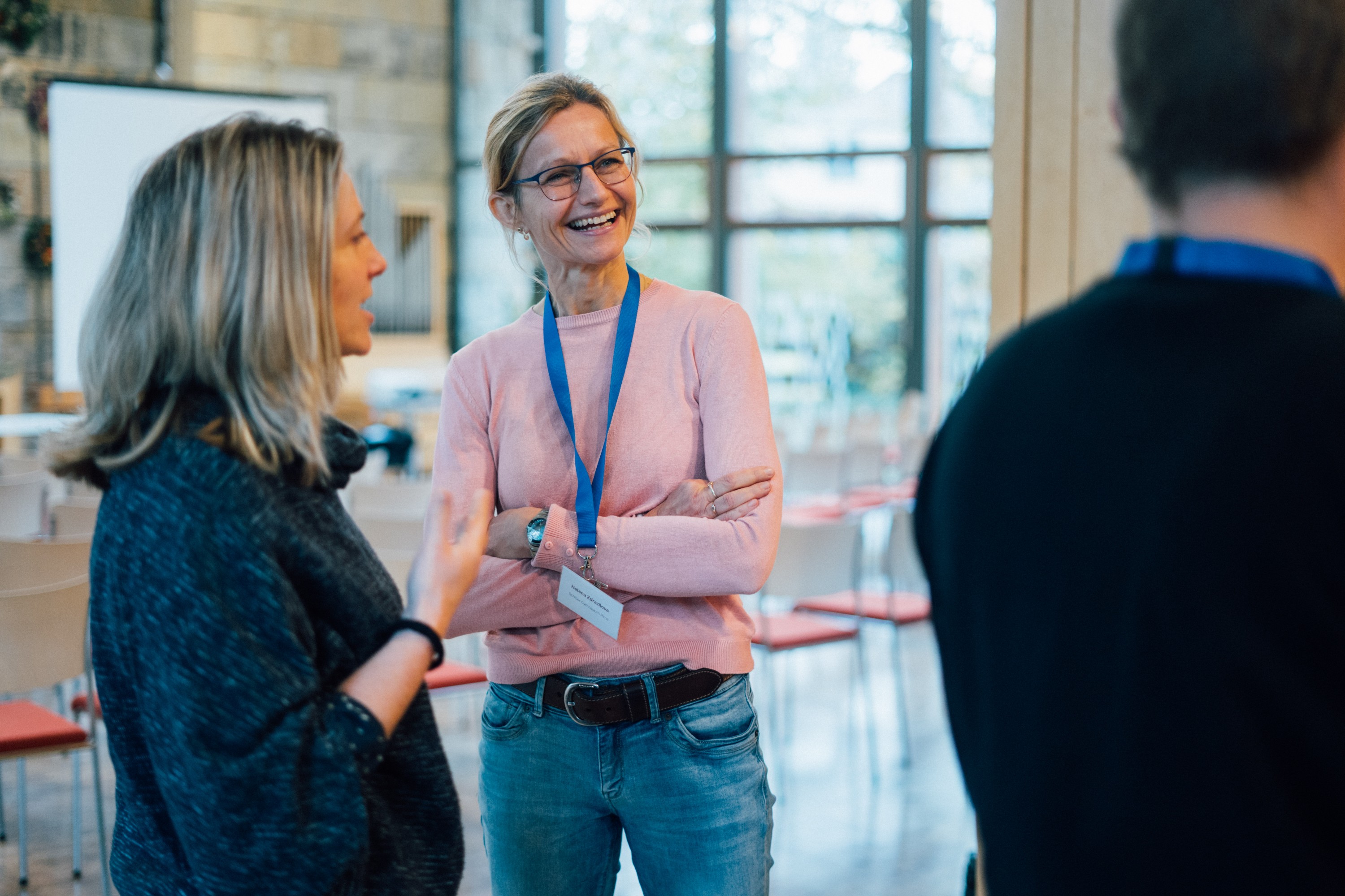 KI generiert: Das Bild zeigt drei Personen in einem hellen Raum mit großen Fenstern, die sich angeregt unterhalten. Die Person in der Mitte trägt ein Namensschild und ein blaues Lanyard, was auf eine Konferenz oder ein Meeting hindeutet.