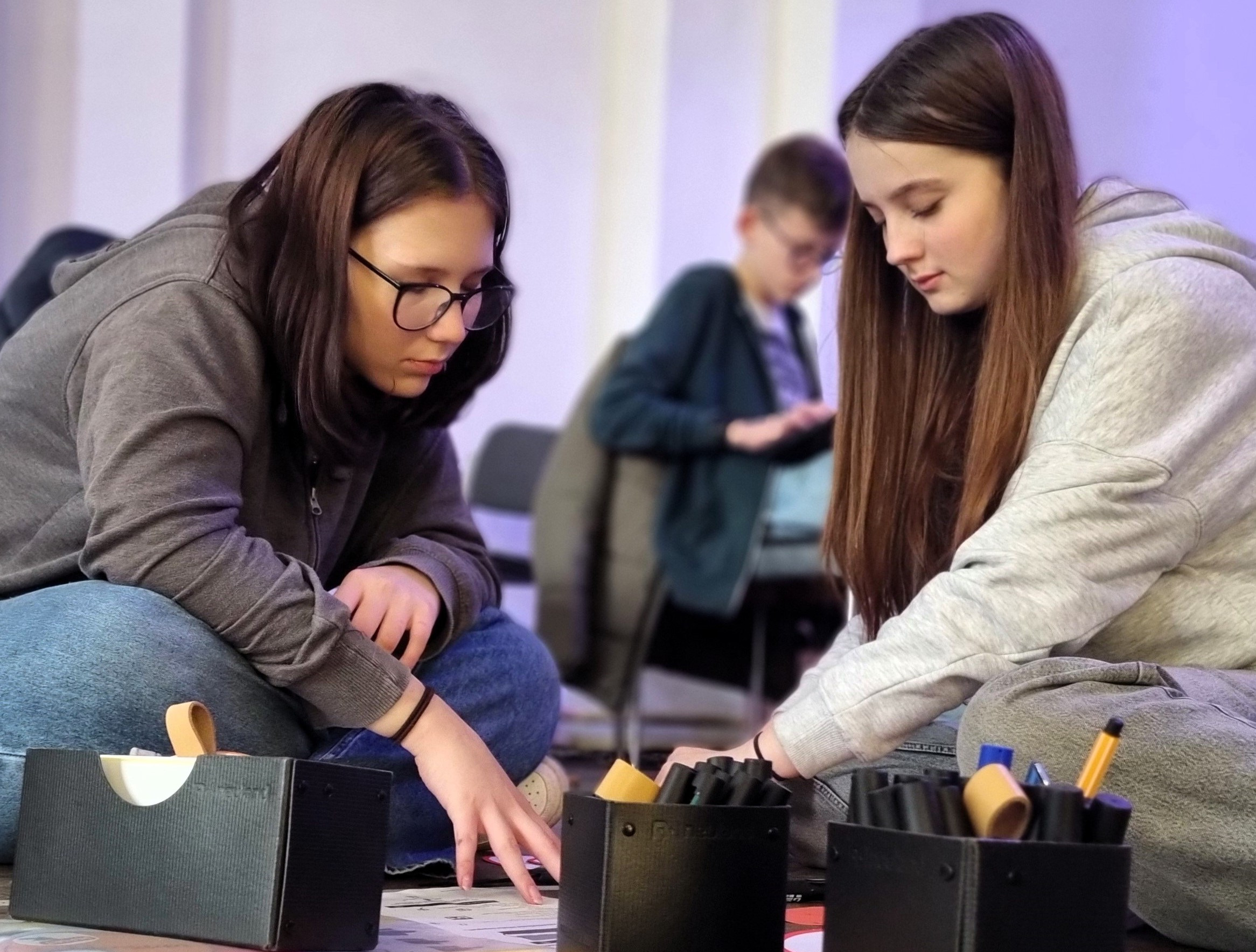 KI generiert: Das Bild zeigt zwei junge Frauen, die sich über einen Tisch beugen und gemeinsam an einem Projekt arbeiten, bei dem sie Materialien aus mehreren Organizer-Behältern verwenden. Im Hintergrund sitzt eine weitere Person, die ebenfalls an einer ähnlichen Aufgabe arbeitet.