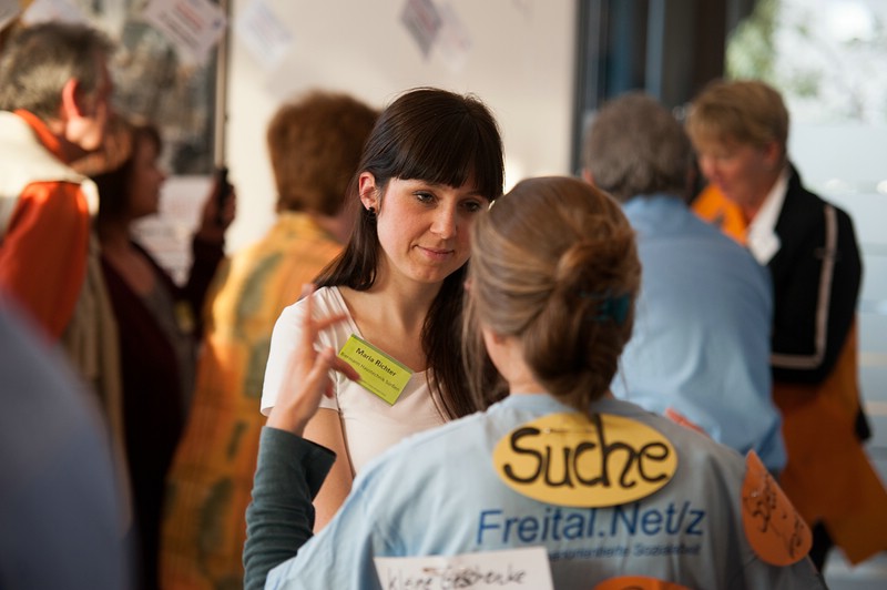 KI generiert: Das Bild zeigt eine Gruppe von Personen, die sich in einem Innenraum unterhalten, wobei eine Frau im Vordergrund mit einer anderen Person spricht. Die Hauptaktivität scheint ein informeller Austausch oder eine Diskussion zu sein, möglicherweise im Rahmen einer Veranstaltung.