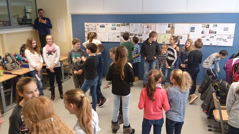 KI generiert: Das Bild zeigt eine Gruppe von Schülern in einem Klassenzimmer, die sich um eine Pinnwand mit verschiedenen Postern und Präsentationen versammelt haben. Der Hauptinhalt des Bildes ist eine Schulpräsentation oder ein Projekt, bei dem die Schüler ihre Arbeiten betrachten und diskutieren.