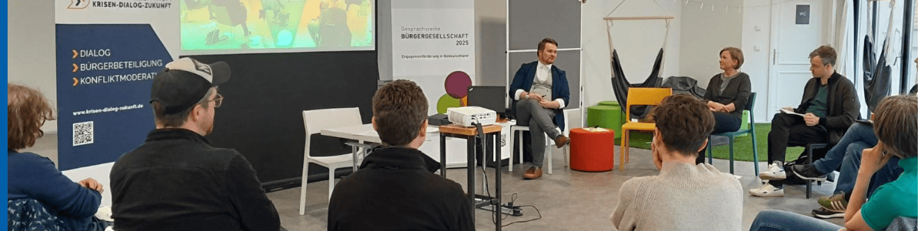 KI generiert: Das Bild zeigt eine Gruppe von Personen in einem Seminarraum, wobei drei Personen auf Stühlen auf einem Podium sitzen und eine Diskussion oder Präsentation führen. Im Hintergrund ist eine Leinwand mit einer Präsentation und ein Banner mit der Aufschrift "Bürgergespräch 2019" zu sehen.