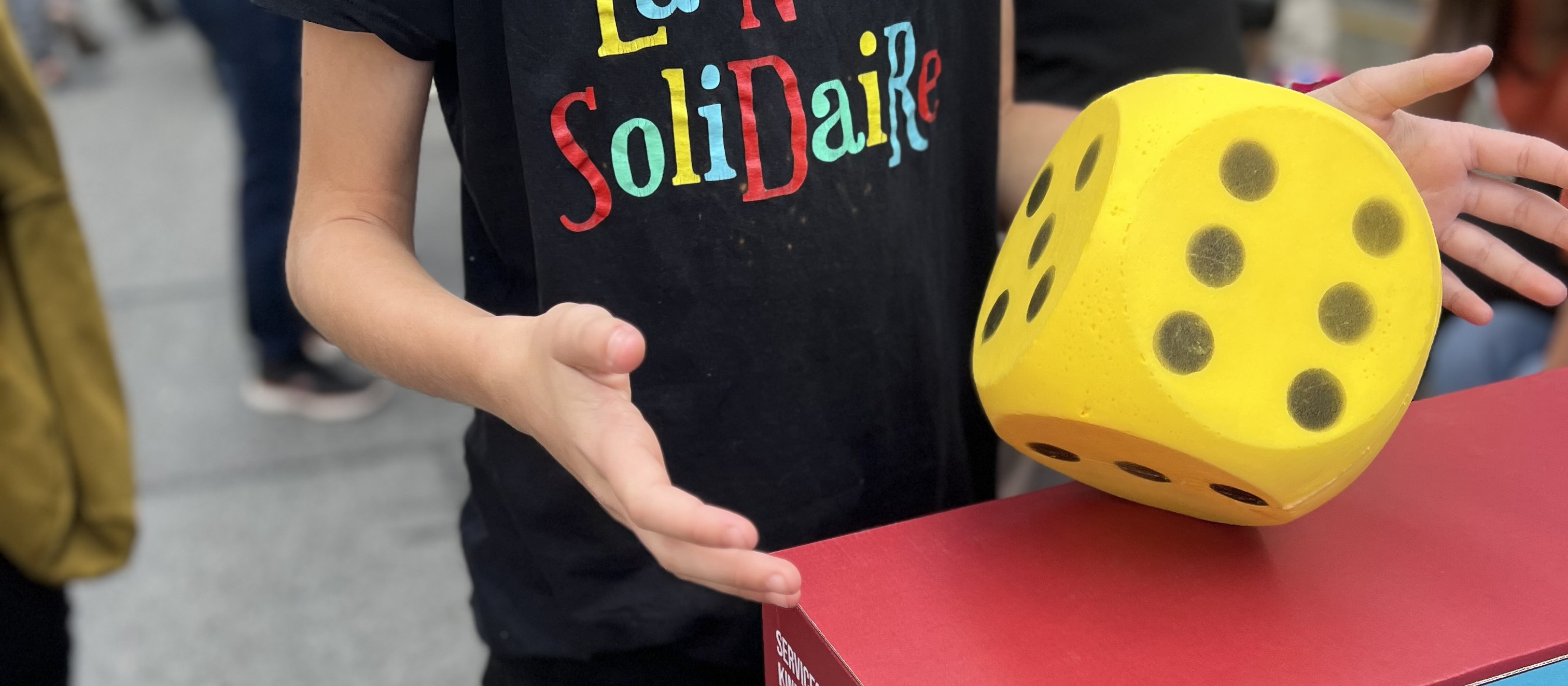 KI generiert: Das Bild zeigt eine Person, die ein großes gelbes Würfelspiel in der Hand hält, während eine andere Person ihre Hand ausstreckt, um den Würfel zu greifen. Im Vordergrund ist ein roter Tisch mit der Aufschrift "SoliDaiRe" zu sehen.