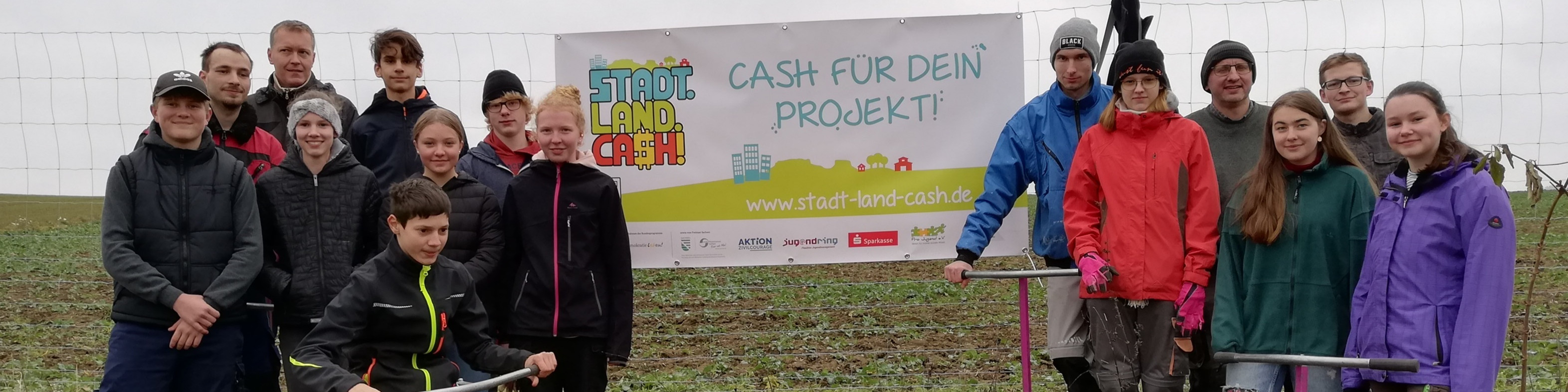 KI generiert: Das Bild zeigt eine Gruppe von Personen, die vor einem Banner mit der Aufschrift "CASH FÜR DEIN PROJEKT!" und der Website "www.stadt-land-cash.de" posieren. Der Hauptinhalt des Bildes ist die Präsentation oder Unterstützung eines Projekts oder Wettbewerbs, der durch das Banner beworben wird.