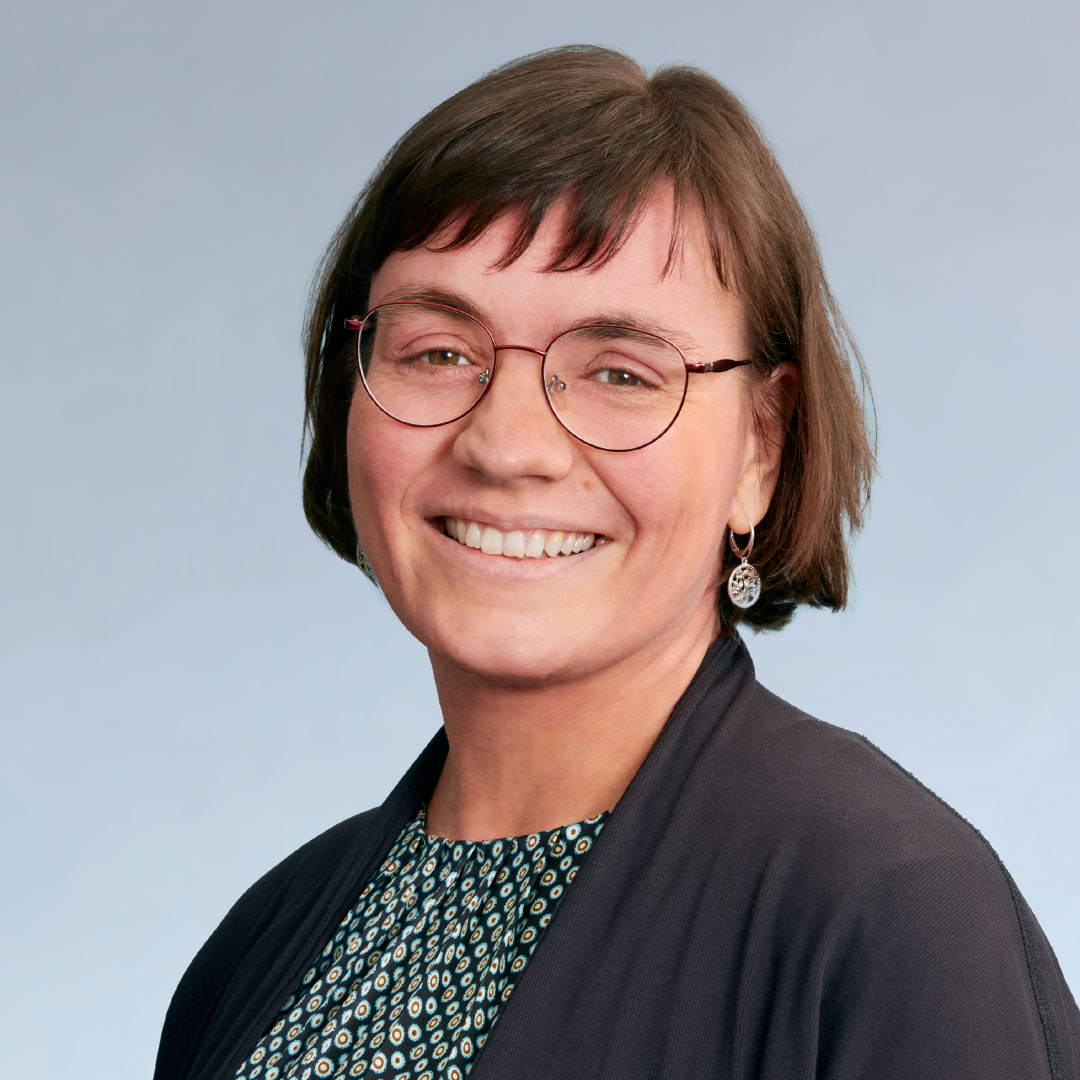 KI generiert: Das Bild zeigt eine lächelnde Frau mit kurzen braunen Haaren und einer Brille. Sie trägt ein dunkles Oberteil über einem gemusterten Kleid und hat kleine, runde Ohrringe.