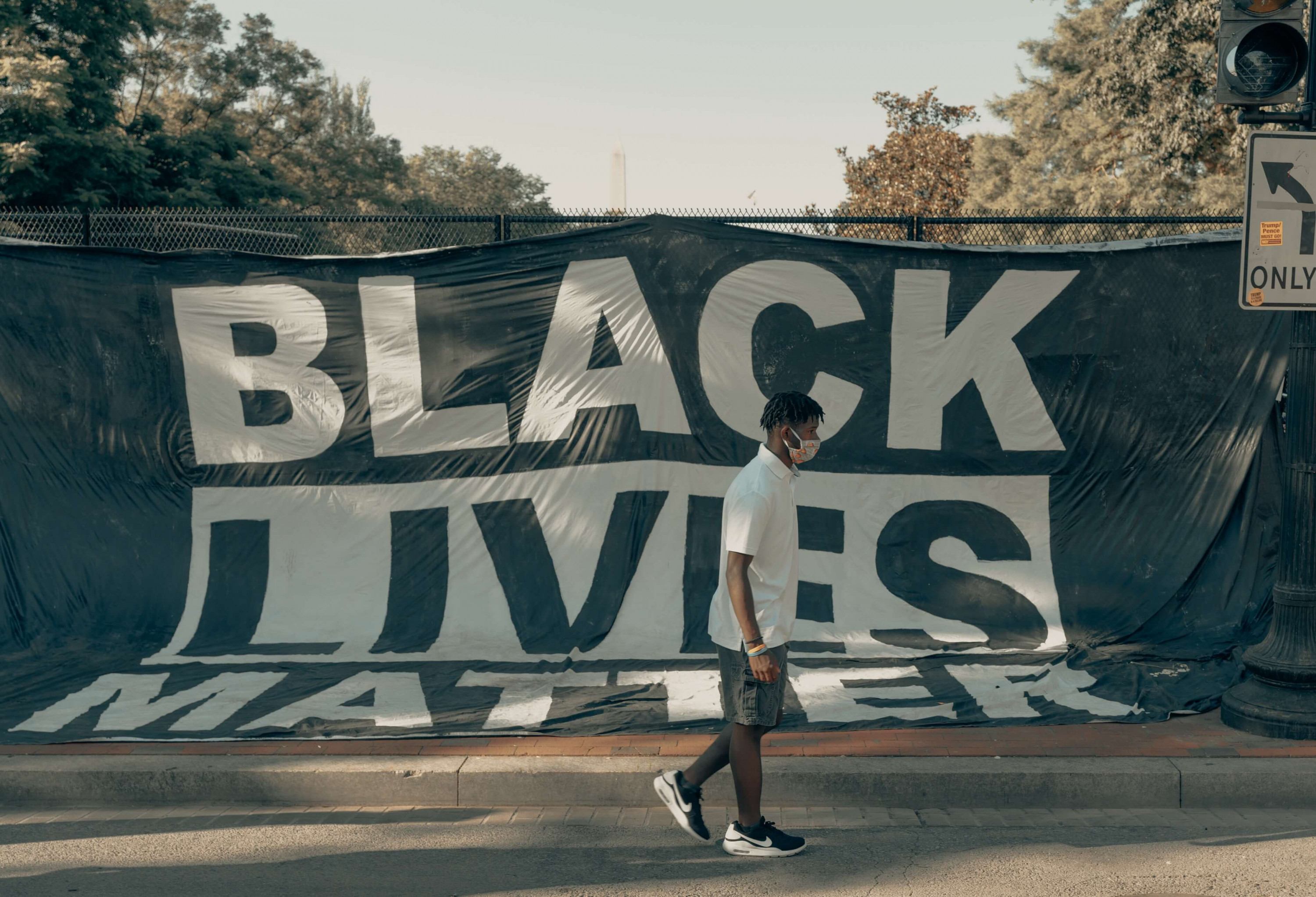 KI generiert: Das Bild zeigt eine Person, die an einem großen Banner mit der Aufschrift "BLACK LIVES MATTER" vorbeigeht. Das Banner ist an einem Zaun befestigt, und im Hintergrund sind Bäume und ein Verkehrsschild zu sehen.
