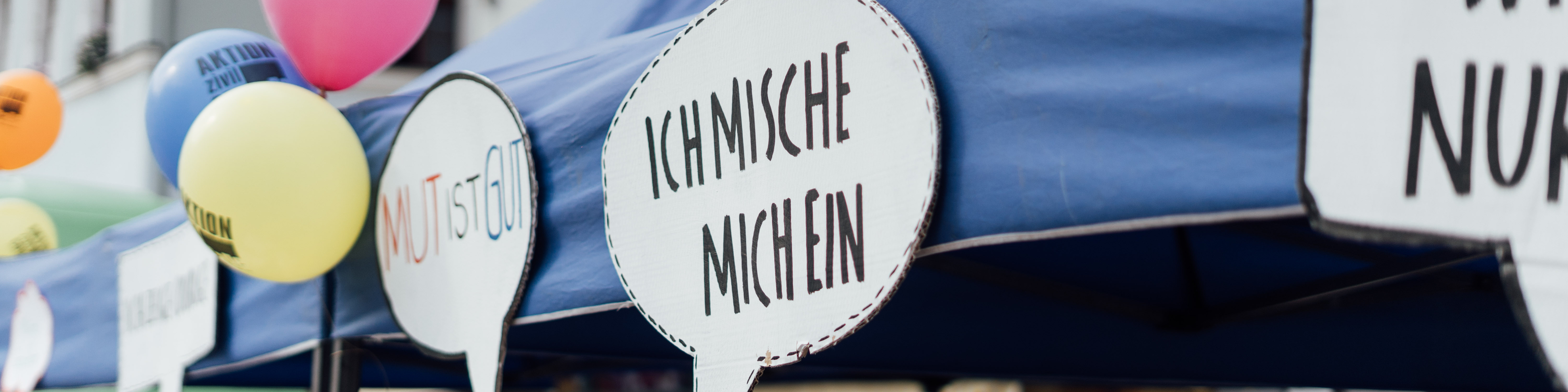 KI generiert: Das Bild zeigt eine Reihe von Sprechblasen mit deutschen Aufschriften, darunter "ICH MISCHE MICH EIN" und "MUT IST GUT", die an einem blauen Zelt oder einer blauen Plane befestigt sind. Im Hintergrund sind bunte Ballons zu sehen, was auf eine Veranstaltung oder ein Fest hindeutet.