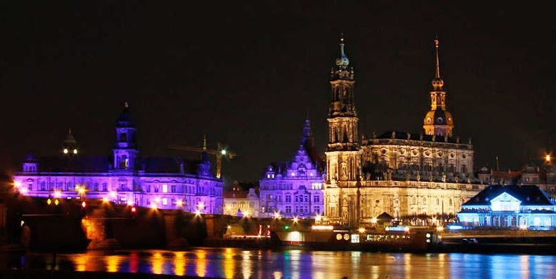 KI generiert: Das Bild zeigt eine nächtliche Stadtansicht mit beleuchteten historischen Gebäuden, darunter eine große Kathedrale und ein Schloss, die sich am Ufer eines Flusses befinden. Der Hauptinhalt des Bildes ist die beeindruckende Architektur und die farbenfrohe Beleuchtung der Gebäude.