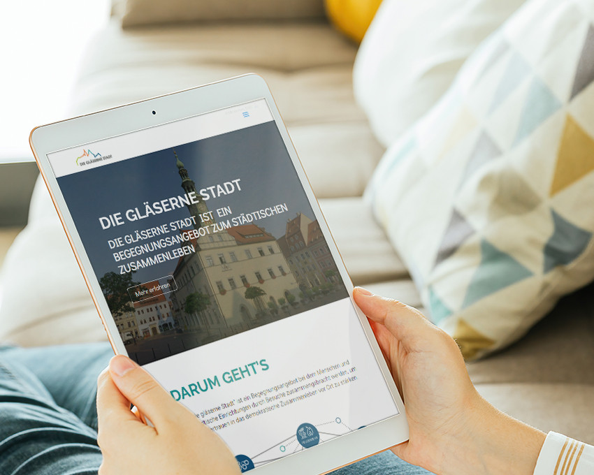 KI generiert: Das Bild zeigt ein Tablet, das eine Webseite mit dem Titel "DIE GLÄSERNE STADT" anzeigt, was auf ein Projekt oder eine Initiative hinweist, die sich mit dem urbanen Leben und der Begegnung in der Stadt befasst. Im Hintergrund sind ein gemütliches Wohnzimmer mit einem Sofa und einer Decke zu sehen.