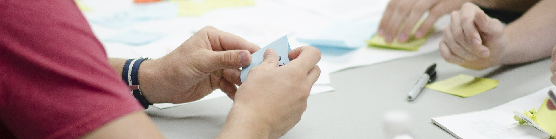 KI generiert: Das Bild zeigt mehrere Personen, die an einem Tisch sitzen und mit bunten Notizzetteln arbeiten, wobei eine Person einen blauen Zettel in der Hand hält. Der Hauptinhalt des Bildes ist eine Gruppenarbeit oder ein Brainstorming, bei dem Ideen auf Notizzetteln festgehalten und diskutiert werden.