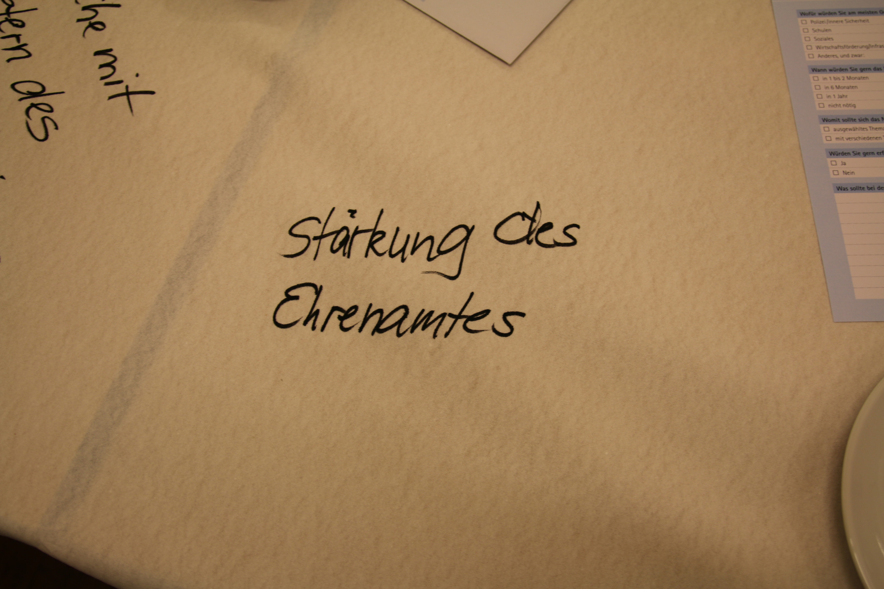 KI generiert: Das Bild zeigt eine Tischdecke mit der handschriftlichen Aufschrift "Stärkung des Ehrenamtes". Im Hintergrund sind weitere handschriftliche Notizen und ein Blatt Papier zu erkennen.