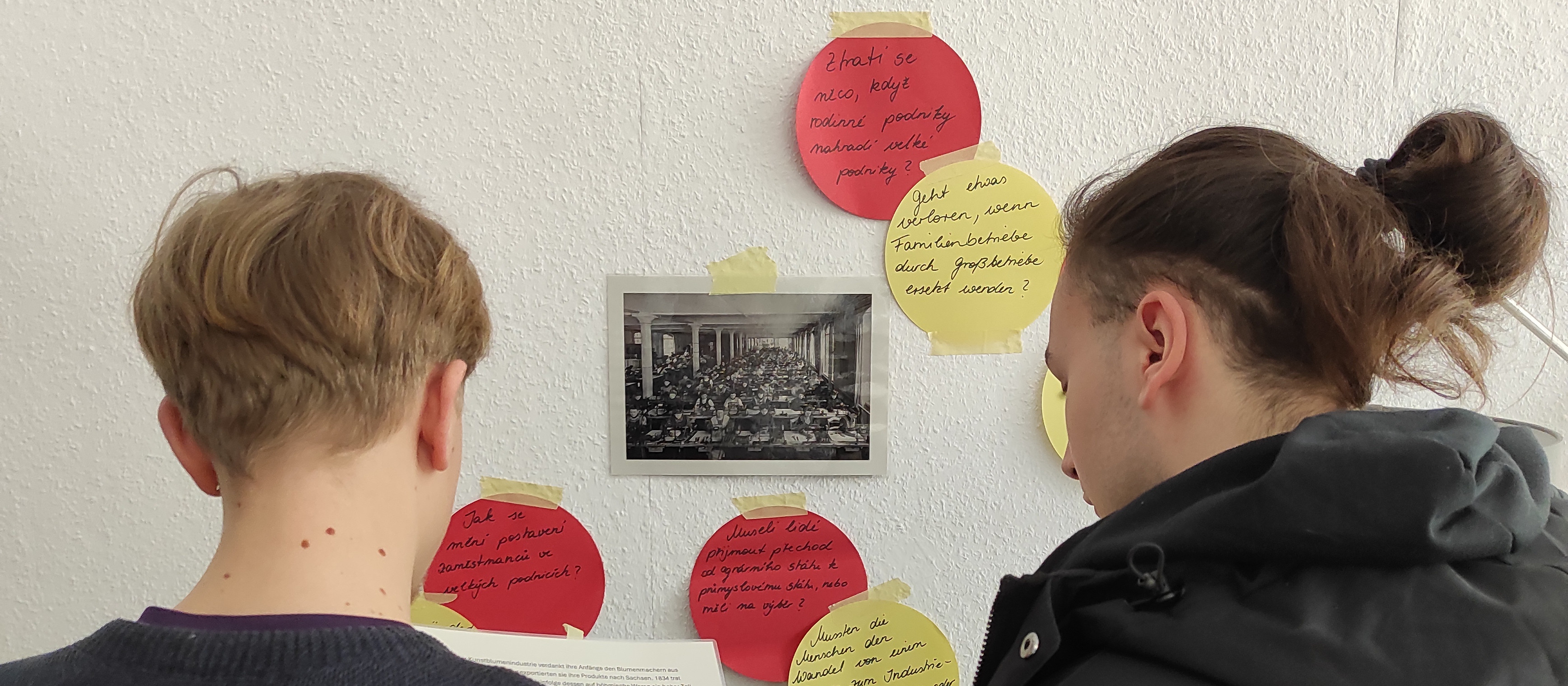 KI generiert: Das Bild zeigt zwei Personen von hinten, die eine Collage an einer Wand betrachten, die aus einem zentralen historischen Foto und mehreren farbigen Notizzetteln mit handschriftlichen Notizen besteht. Der Hauptinhalt des Bildes ist die Interaktion der Personen mit der Collage, die möglicherweise eine Diskussion oder Reflexion über das dargestellte Thema anregt.