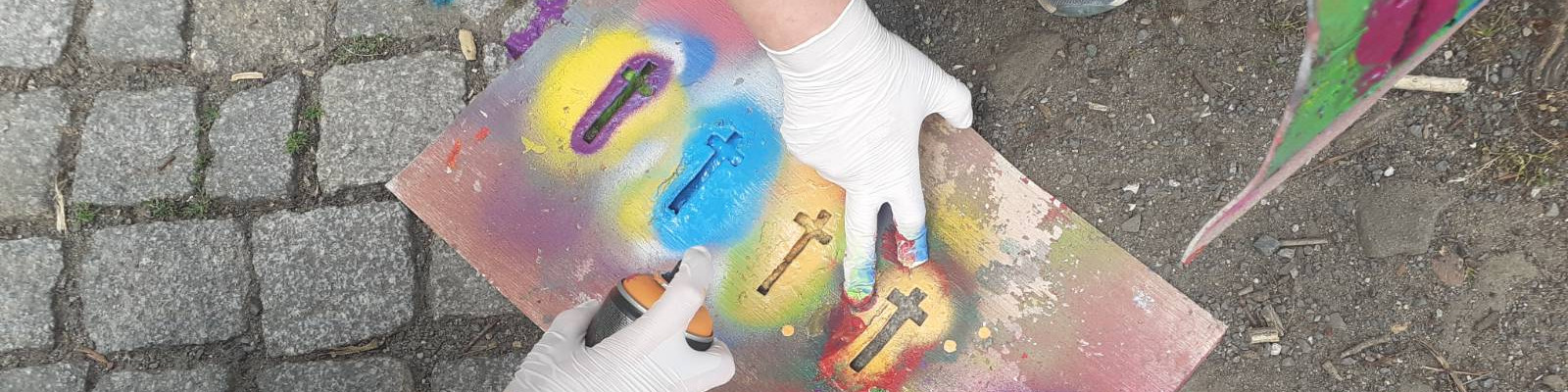 KI generiert: Das Bild zeigt eine Person, die mit Sprühdosen und Schablonen auf einem Stück Pappe Graffiti sprüht. Der Hauptinhalt ist das Erstellen von Street-Art mit religiösen Symbolen wie Kreuzen.