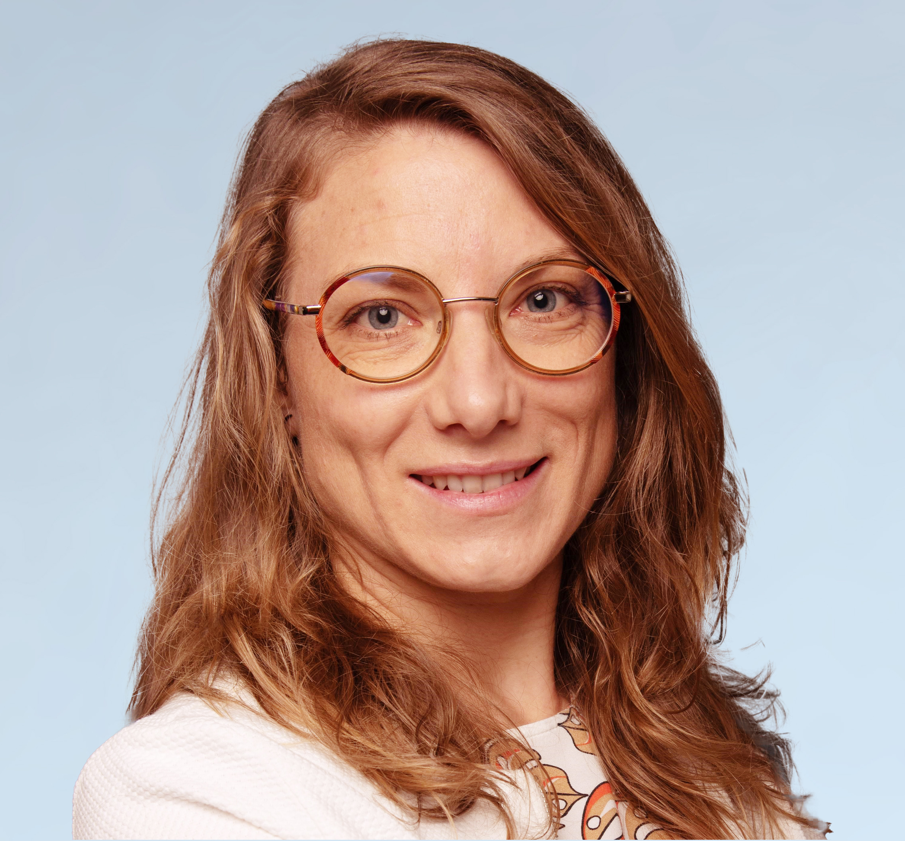 KI generiert: Das Bild zeigt eine lächelnde Frau mit langen, welligen, braunen Haaren und runden Brillen mit braunem Rahmen. Sie trägt ein helles Oberteil mit einem Muster und hat einen freundlichen Gesichtsausdruck.