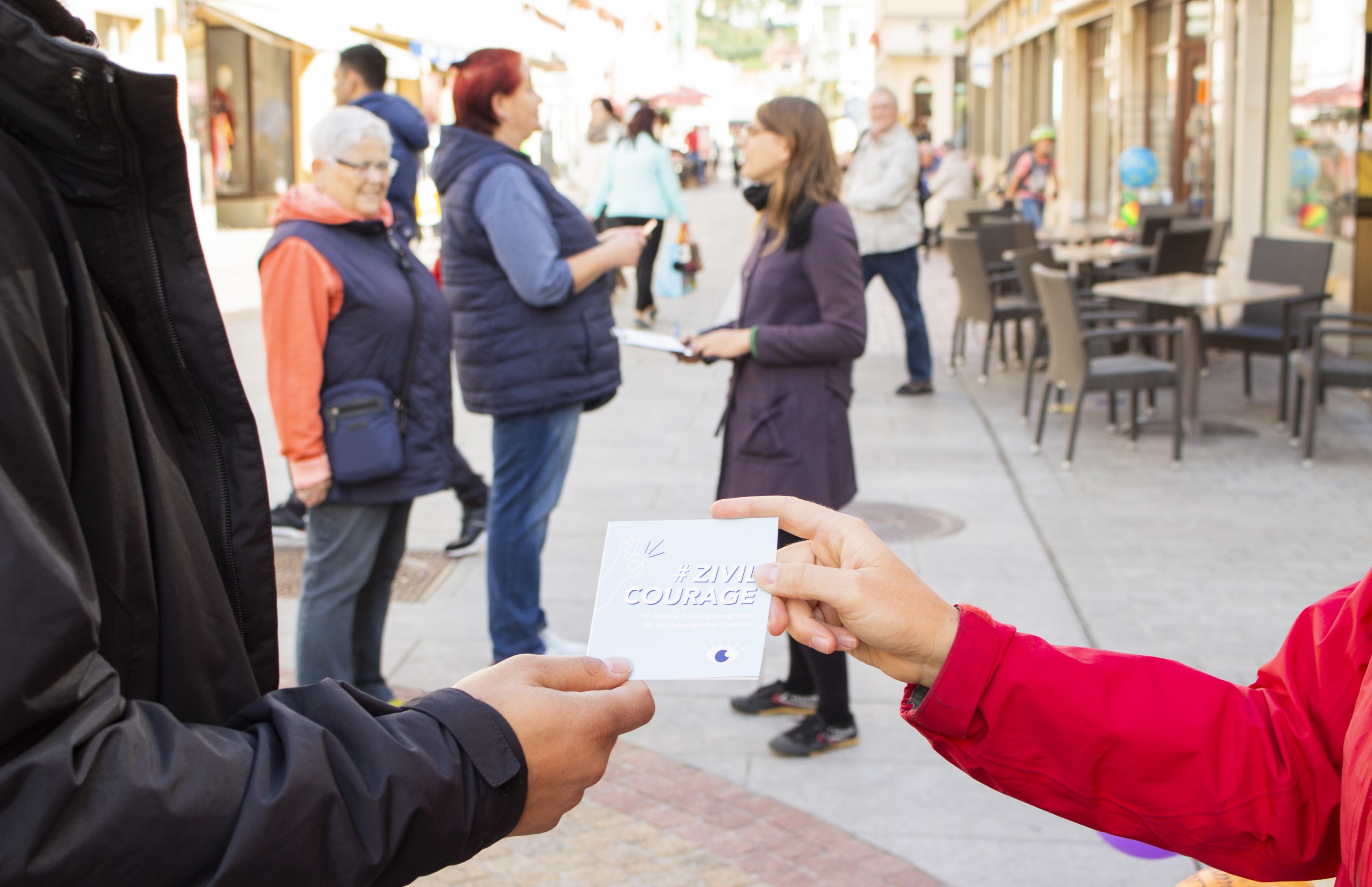 KI generiert: Das Bild zeigt eine Person in einem roten Mantel, die einer anderen Person ein Stück Papier mit der Aufschrift "#ZIVILCOURAGE" überreicht. Im Hintergrund sind mehrere Menschen auf einer belebten Straße zu sehen, einige von ihnen scheinen sich zu unterhalten.
