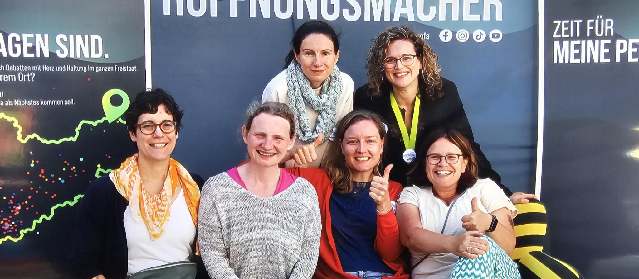 KI generiert: Das Bild zeigt eine Gruppe von sieben lächelnden Frauen, die eng beieinander sitzen und in die Kamera schauen. Im Hintergrund ist ein Werbeplakat mit dem Text "HOFFNUNGSMACHER" und "ZEIT FÜR MEINE PERS" zu sehen.