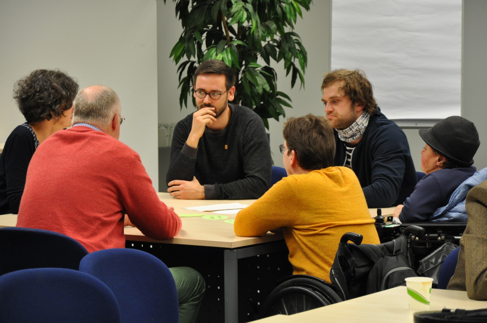 KI generiert: Das Bild zeigt eine Gruppe von Personen, die an einem Tisch sitzen und sich in einer Besprechung oder Diskussion befinden. Der Hauptinhalt des Bildes ist eine Meeting-Situation in einem Büro oder Besprechungsraum.