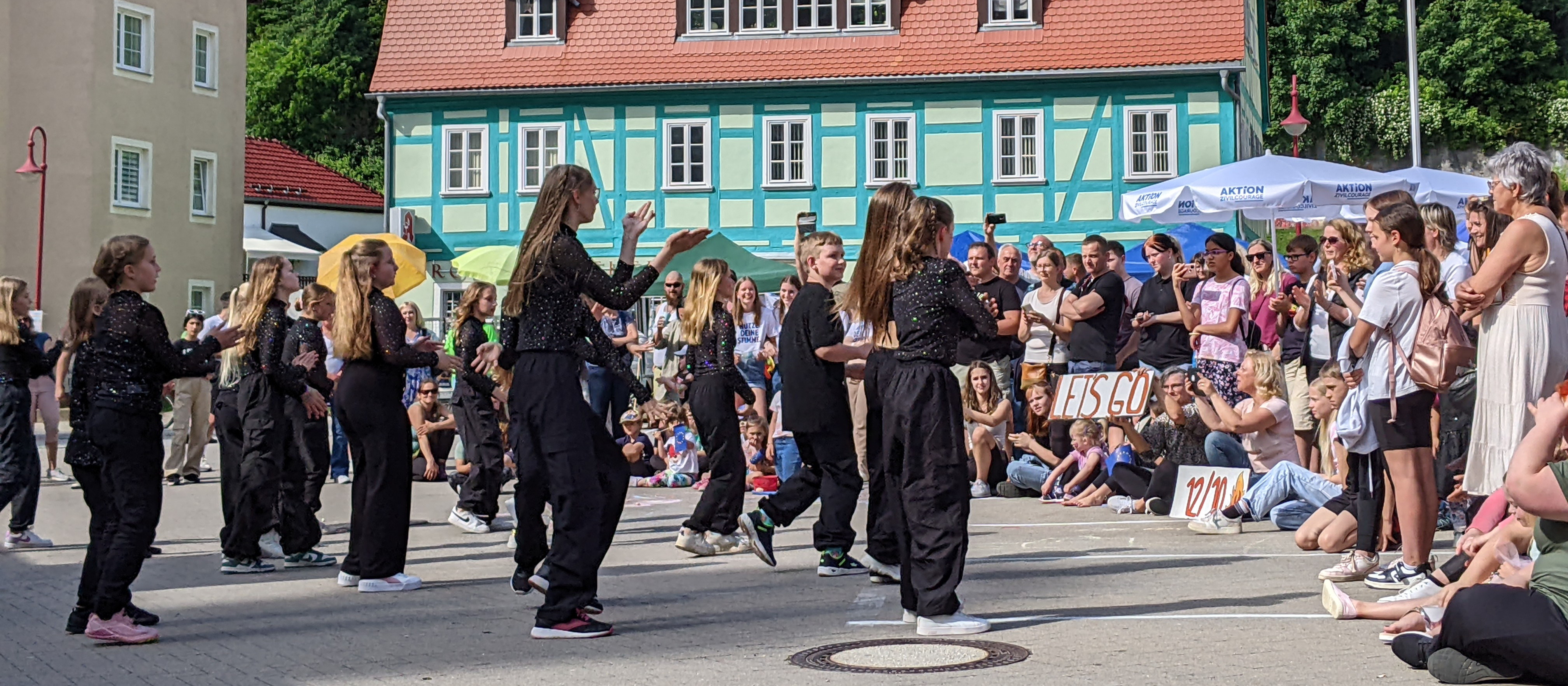 KI generiert: Das Bild zeigt eine Gruppe von Personen, die auf einer Straße tanzen, während eine Menschenmenge zuschaut. Im Hintergrund sind bunte Gebäude und ein Zelt zu sehen.