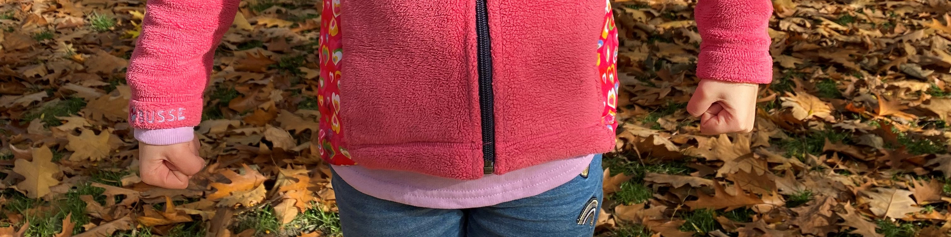 KI generiert: Das Bild zeigt den Oberkörper und die Arme eines Kindes, das eine pinkfarbene Jacke mit bunten Mustern auf den Ärmeln trägt und auf einem Boden voller herbstlicher Blätter steht. Der Hauptinhalt des Bildes ist das Kind in der Outdoor-Umgebung.