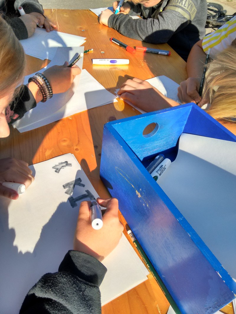 KI generiert: Das Bild zeigt eine Gruppe von Personen, die an einem Holztisch sitzen und auf Papier zeichnen oder schreiben. Der Hauptinhalt des Bildes ist eine kreative oder künstlerische Aktivität, bei der verschiedene Werkzeuge wie Stifte, Marker und ein blauer Behälter mit Materialien verwendet werden.