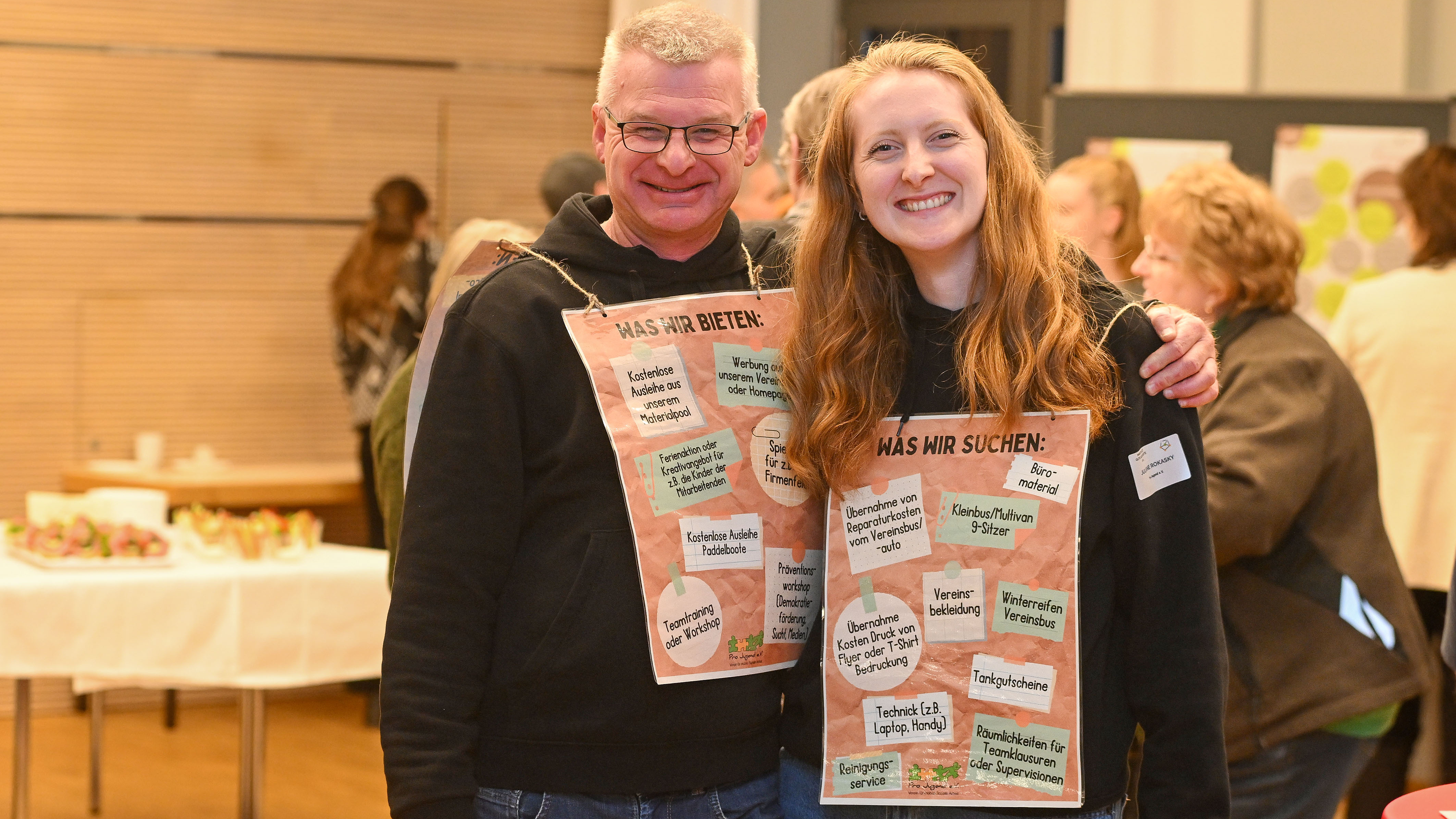 KI generiert: Das Bild zeigt zwei lächelnde Personen, die jeweils ein Plakat mit Aufschriften halten, die sich auf das Thema "Was wir bieten" und "Was wir suchen" beziehen. Im Hintergrund sind weitere Personen und ein Tisch mit Speisen zu erkennen, was auf eine Veranstaltung oder ein Treffen hindeutet.