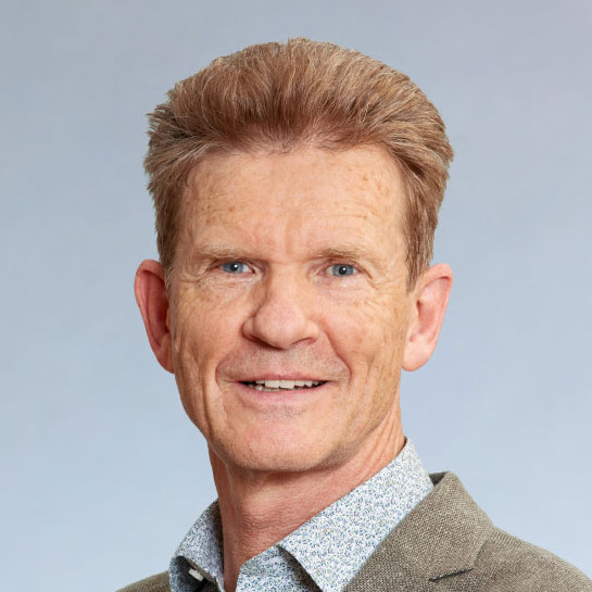 KI generiert: Das Bild zeigt ein Porträt eines lächelnden Mannes mit kurzen, rötlichen Haaren und blauen Augen. Er trägt ein helles Hemd mit einem Muster und eine beige Jacke.