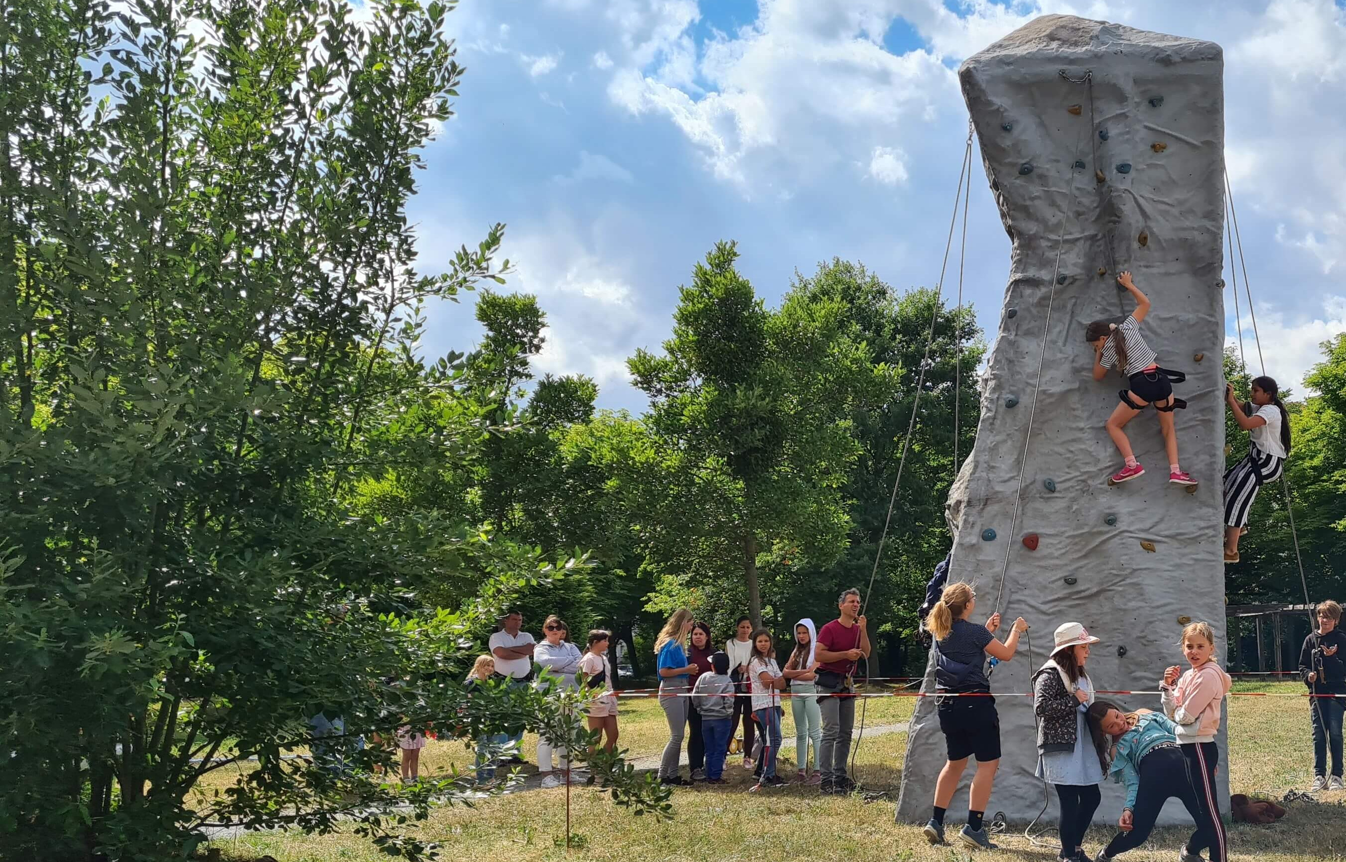 KI generiert: Das Bild zeigt eine Gruppe von Menschen, die eine Kletterwand im Freien nutzen, wobei eine Person gerade klettert und andere zuschauen oder warten. Der Hauptinhalt des Bildes ist die Kletterwand und die Aktivität des Kletterns in einer natürlichen Umgebung.