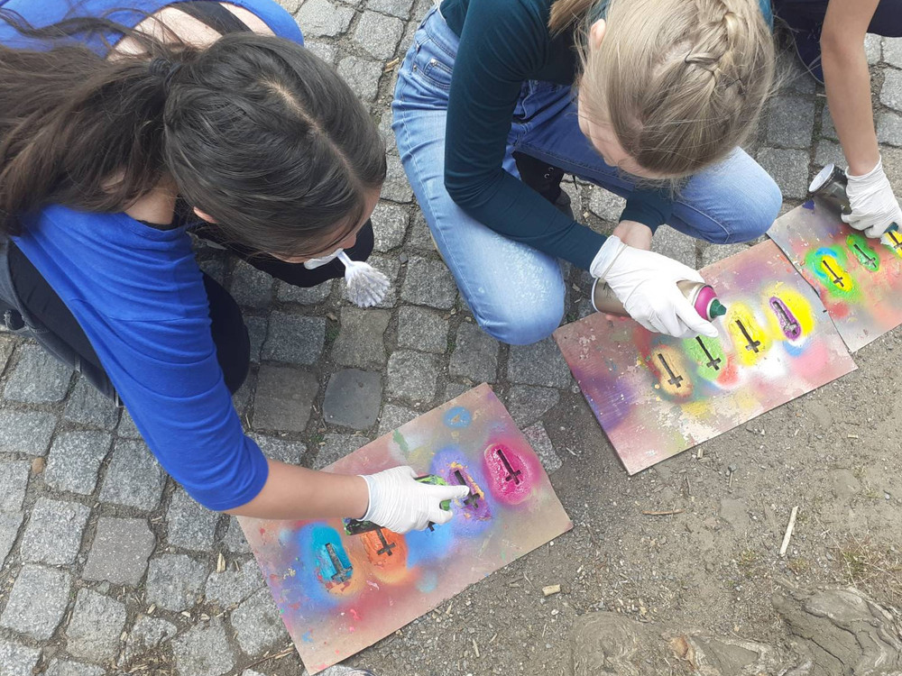 KI generiert: Das Bild zeigt eine Person, die mit Sprühdosen und Schablonen auf einem Stück Pappe Graffiti sprüht. Der Hauptinhalt ist das Erstellen von Street-Art mit religiösen Symbolen wie Kreuzen.