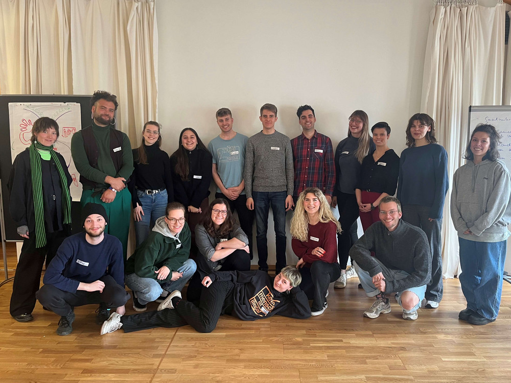 KI generiert: Das Bild zeigt eine Gruppe von Personen, die in einem hellen Raum mit Holzfußboden sitzen und einer Präsentation oder einem Vortrag folgen. Im Vordergrund sind Zuhörer zu sehen, während im Hintergrund eine Person vor einem Projektor steht und eine Präsentation hält.