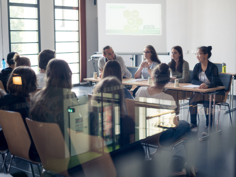 KI generiert: Das Bild zeigt eine Gruppe von Personen, die in einem hellen Raum an Tischen sitzen und sich in einer Diskussion oder einem Meeting befinden. Der Hauptinhalt des Bildes ist eine Besprechung oder ein Workshop in einem professionellen oder akademischen Umfeld.
