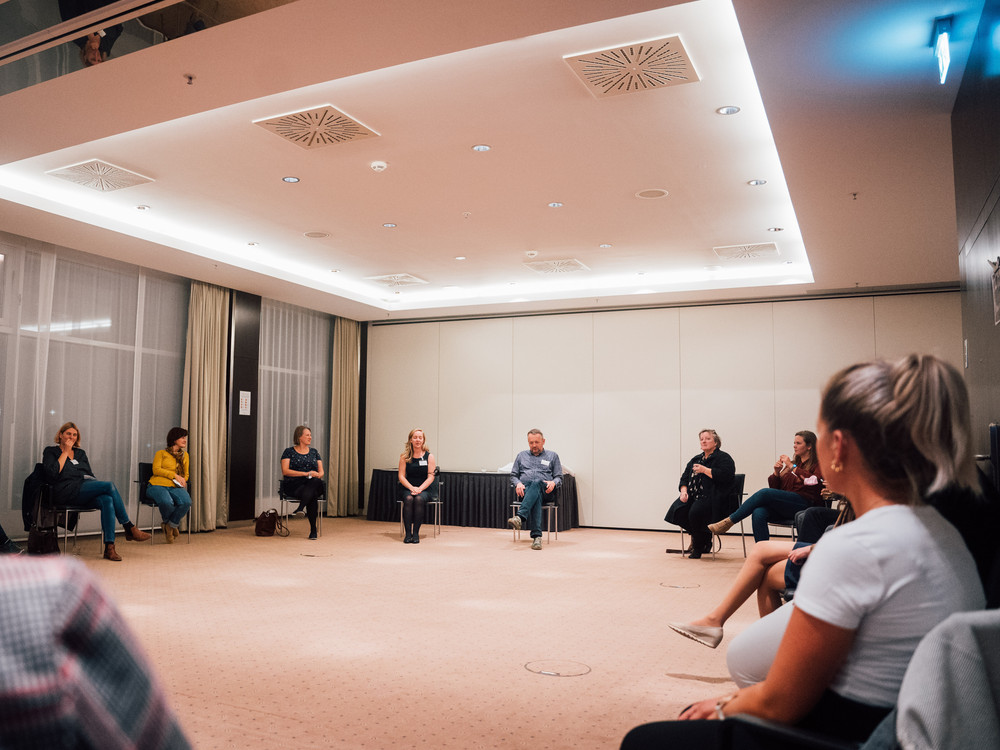 KI generiert: Das Bild zeigt eine Gruppe von Personen, die in einem hellen Raum auf Stühlen im Kreis sitzen und sich unterhalten. Der Hauptinhalt ist eine Diskussion oder ein Meeting in einer informellen Atmosphäre.