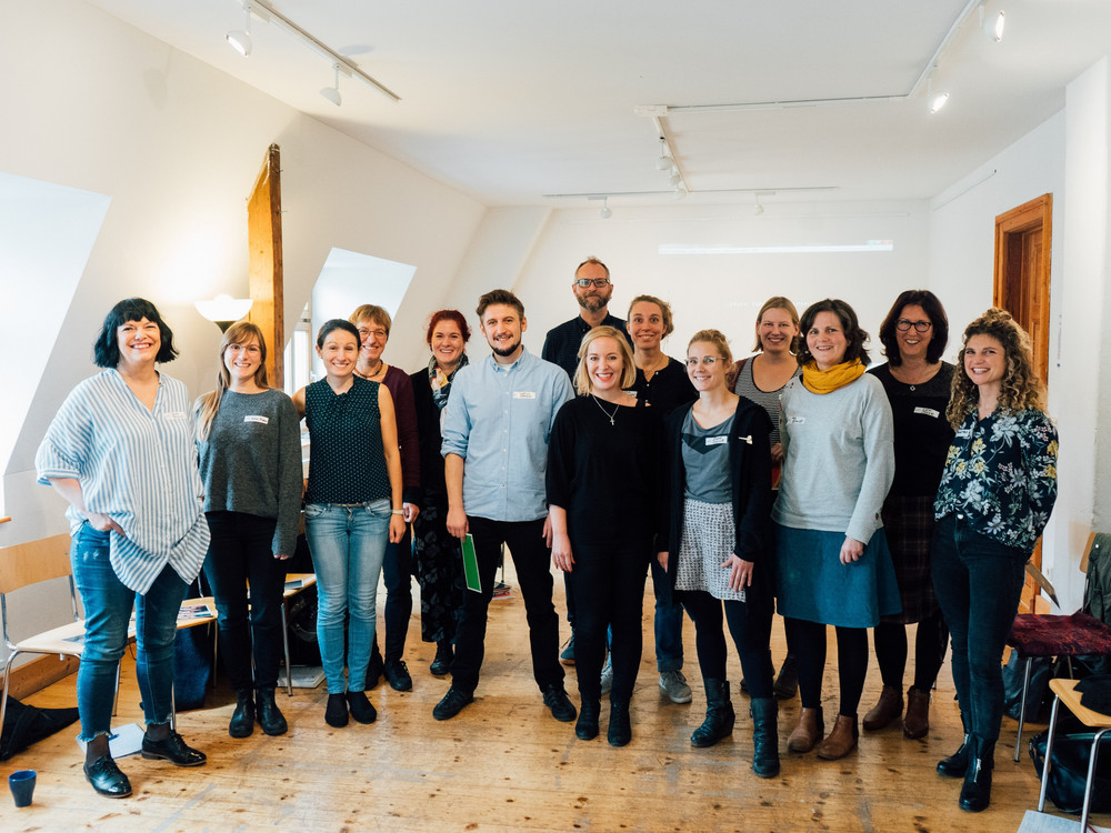 KI generiert: Das Bild zeigt eine Gruppe von Personen, die in einem hellen Raum mit Holzboden und schrägen Wänden stehen und für ein Gruppenfoto posieren. Der Hauptinhalt des Bildes ist die Gruppe von Menschen, die gemeinsam ein Foto machen.