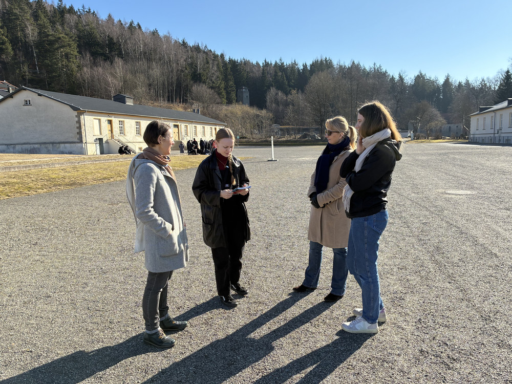 KI generiert: Das Bild zeigt eine Gruppe von vier Personen, die sich auf einem großen, offenen Platz unterhalten, während sie auf ein Smartphone schauen. Im Hintergrund sind Gebäude und eine Gruppe von Menschen zu sehen, die möglicherweise eine Führung oder Besichtigung machen.