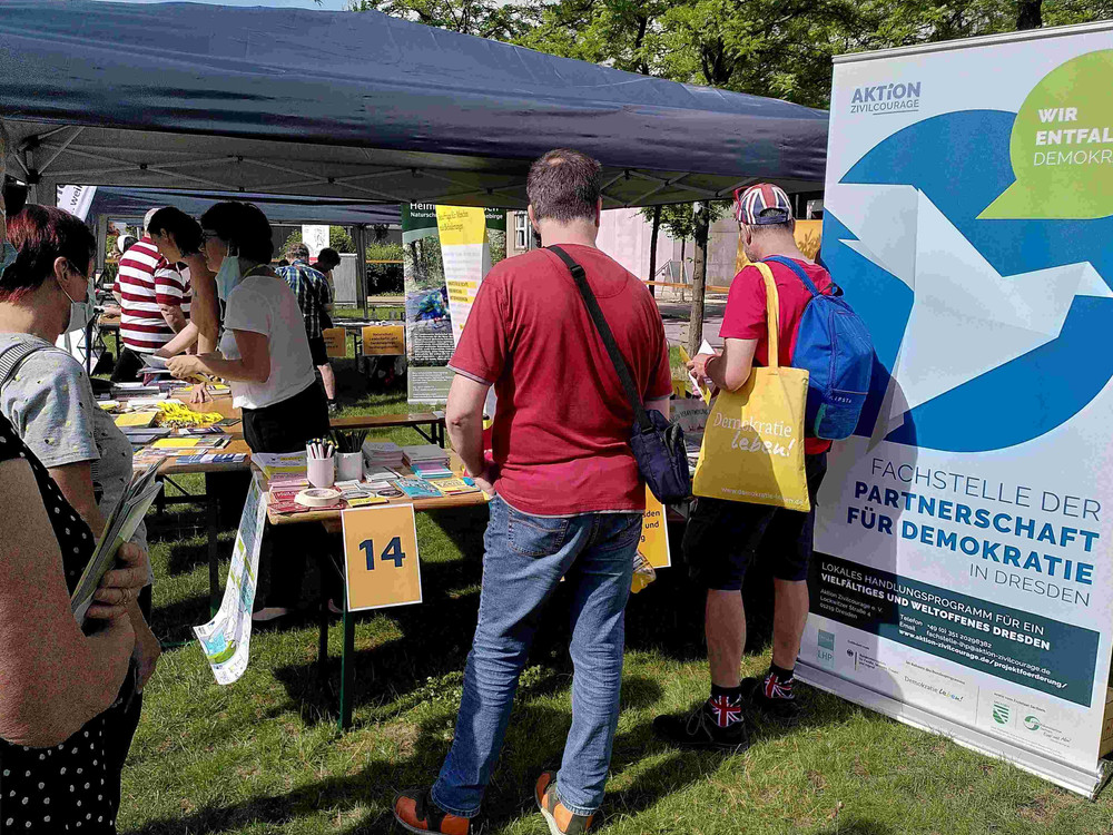 KI generiert: Das Bild zeigt eine Gruppe von Menschen, die sich an einem Stand auf einer Veranstaltung im Freien versammeln. Der Stand hat ein Banner mit der Aufschrift "Wir entfalten Demokratie" und bietet Informationsmaterial an.