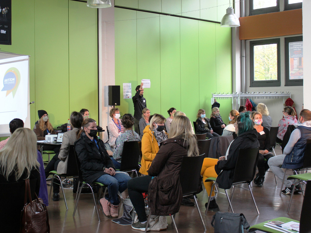 KI generiert: Das Bild zeigt eine Gruppe von Personen, die in einem hellen Raum mit grünen Wänden an einer Veranstaltung teilnehmen. Der Hauptinhalt ist eine Präsentation oder ein Workshop, bei dem eine Person vorne steht und zu den Anwesenden spricht.