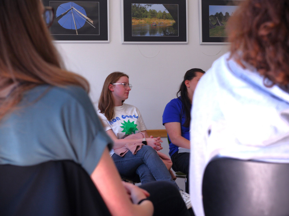 KI generiert: Das Bild zeigt eine Gruppe von Personen, die in einem Raum stehen und sich unterhalten. Der Hauptinhalt ist eine Person mit Brille und einem T-Shirt mit einem Aufdruck, die im Mittelpunkt der Gruppe steht und sich mit anderen unterhält.