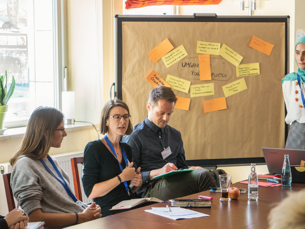 KI generiert: Das Bild zeigt eine Gruppe von Personen in einem Besprechungsraum, die an einem Tisch sitzen und an einer Diskussion oder einem Workshop teilnehmen. Eine Person steht und präsentiert Ideen, die auf einem Flipchart mit Post-its notiert sind.