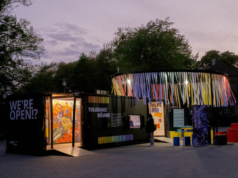 KI generiert: Das Bild zeigt eine moderne, farbenfrohe Installation oder Ausstellung im Freien mit dem Schriftzug "WE'RE OPEN!" und "TOLERANZ RÄUME". Der Hauptinhalt ist eine kreative, interaktive Kunstinstallation, die Passanten einlädt, sie zu erkunden.