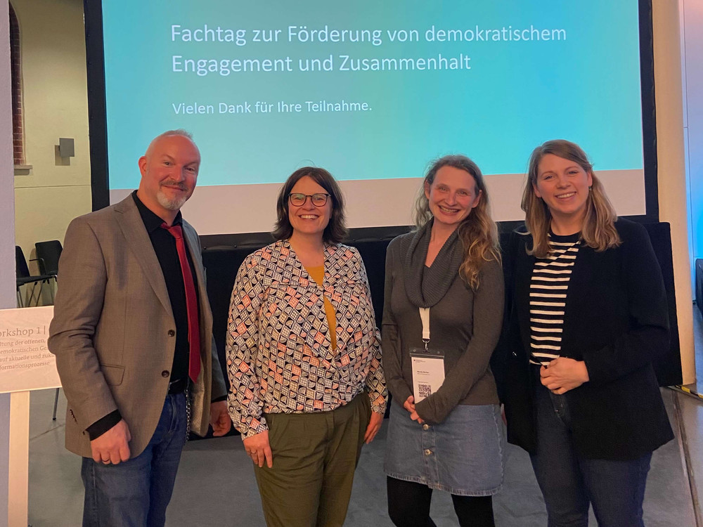 KI generiert: Das Bild zeigt vier lächelnde Personen, die nebeneinander vor einem Projektor oder Bildschirm mit blau-weißen Hintergrund stehen. Der Hauptinhalt des Bildes ist eine Gruppe von Personen, die möglicherweise an einer Präsentation oder einem Meeting teilnehmen.