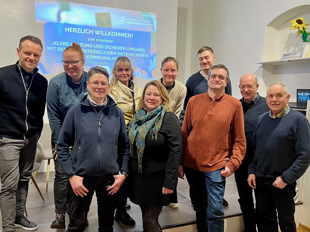 KI generiert: Das Bild zeigt eine Gruppe von zwölf Personen, die gemeinsam in einem Raum posieren. Im Hintergrund ist ein Banner mit Text zu sehen, der auf eine Veranstaltung oder ein Treffen hinweist.