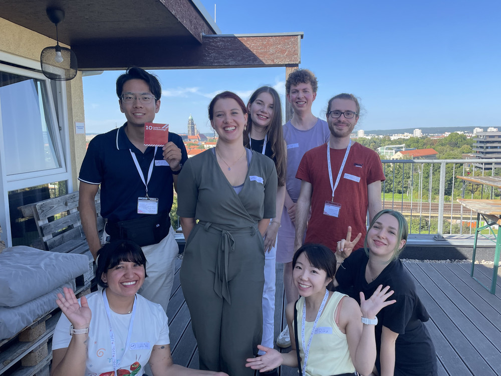 KI generiert: Das Bild zeigt eine Gruppe von fünf Personen, die auf einem Balkon stehen und in die Kamera lächeln. Im Hintergrund ist eine Stadtlandschaft mit Gebäuden und Bäumen zu sehen, und eine Person hält ein rotes Schild mit der Aufschrift "10 Jahre".