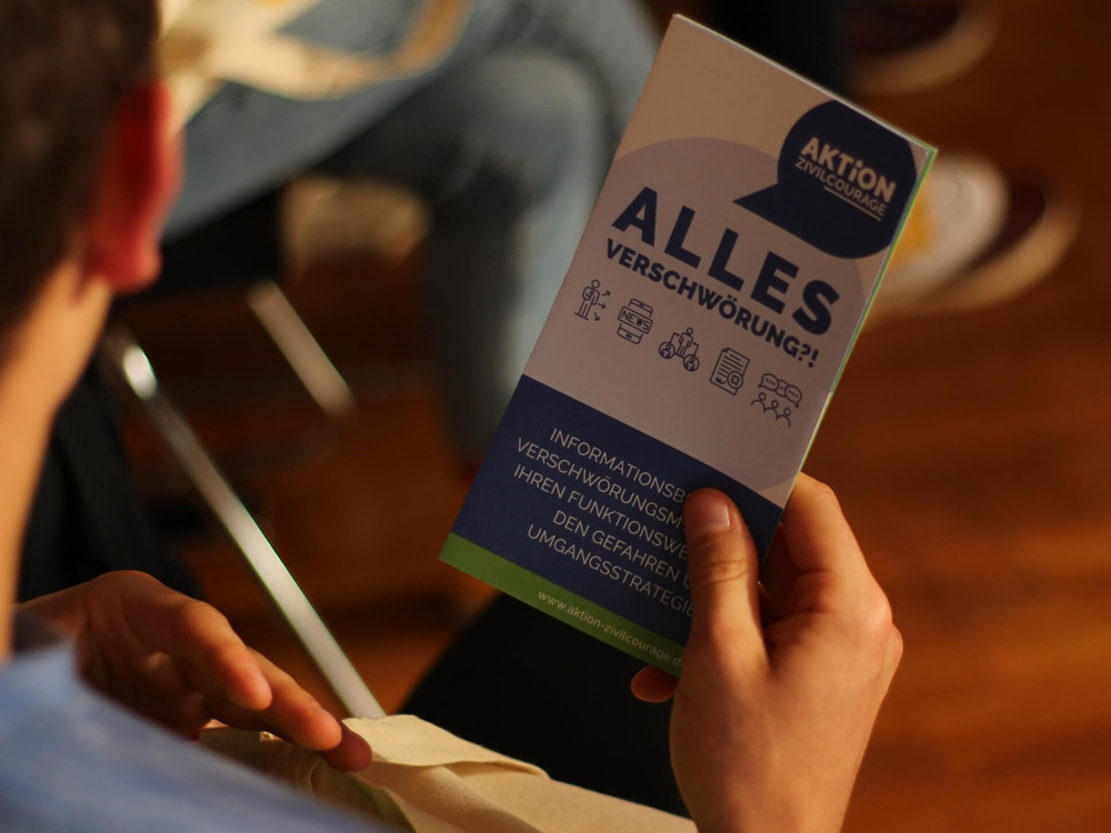 KI generiert: Das Bild zeigt eine Hand, die ein Informationsblatt mit dem Titel "AKTION GRUNDWASSERSCHUTZ" hält, auf dem die Frage "ALLES VERSCHWÖRUNG?!" zu lesen ist. Der Hauptinhalt des Bildes ist das Informationsblatt, das sich mit dem Thema Verschwörungstheorien im Kontext des Grundwasserschutzes beschäftigt.
