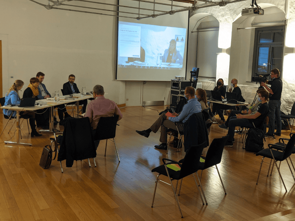 KI generiert: Das Bild zeigt eine Gruppe von Personen in einem Seminarraum, die an langen Tischen sitzen und sich unterhalten oder einer Präsentation folgen. Im Vordergrund sind einige Teilnehmer zu sehen, während im Hintergrund eine Leinwand mit einer Präsentation und ein Kameramann, der die Veranstaltung aufzeichnet, sichtbar sind.