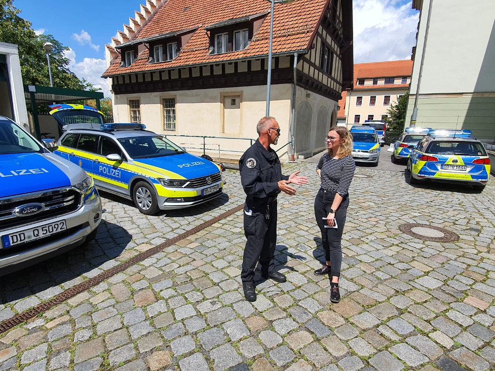 KI generiert: Das Bild zeigt eine Szene auf einem gepflasterten Platz mit mehreren Polizeiwagen im Hintergrund. Im Vordergrund unterhalten sich ein Polizist und eine Frau.